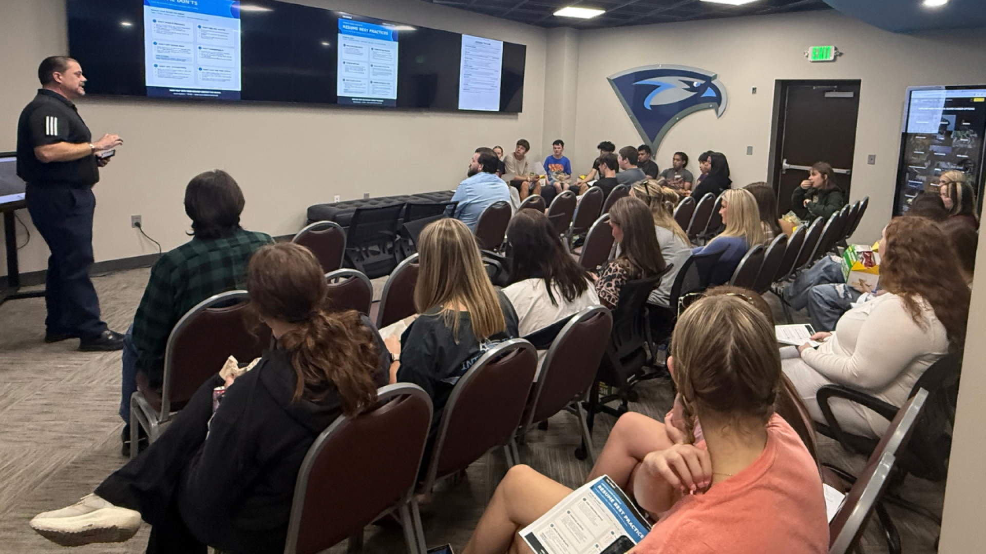 A speaker presents resume best practices on screens to a seated audience of students eating lunch in a room with a hawk mascot logo on the wall.